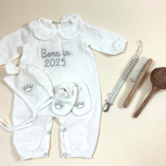 White baby outfit with 'Born in 2025' text, baby booties, and baby care items on a beige background