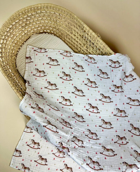 Muslin blanket with rocking horse pattern draped over a wicker basket on a beige background