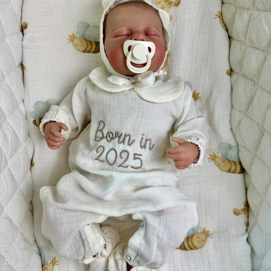 Newborn baby in a white outfit with 'Born in 2025' text, lying on a textured blanket.